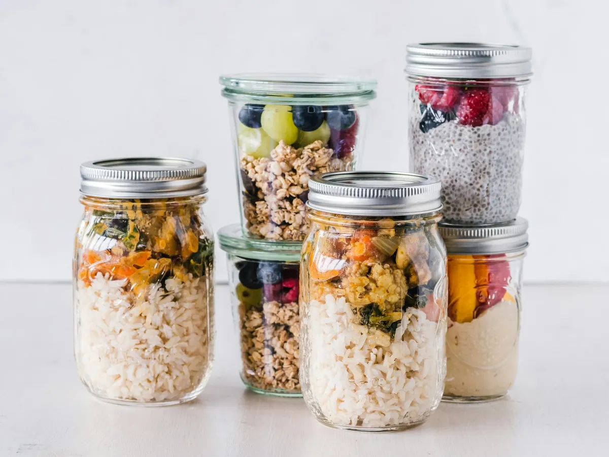 Six glass jars of make-ahead food on a white table: granola with blueberries and grapes, chia pudding topped with raspberries, yogurt under sliced mango and berries, and two jars of rice with roasted vegetables