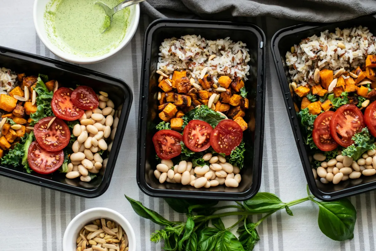 Three black meal prep containers of wild rice, roasted sweet potato, kale, sliced tomatoes and white beans, with a bowl of green dressing, a dish of flaked almonds and a sprig of basil on a striped cloth