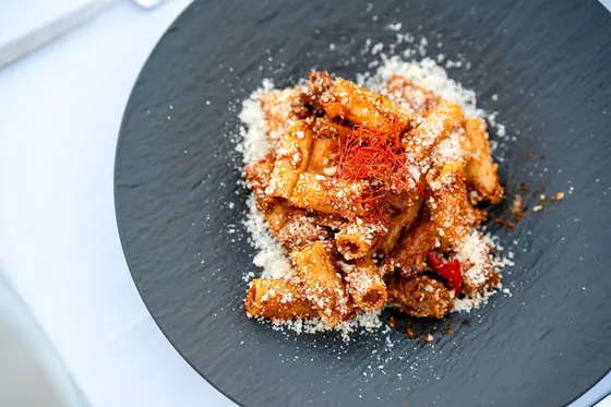 Rigatoni in a meat ragu on a dark slate plate, dusted with grated parmesan and topped with fine threads of red chilli