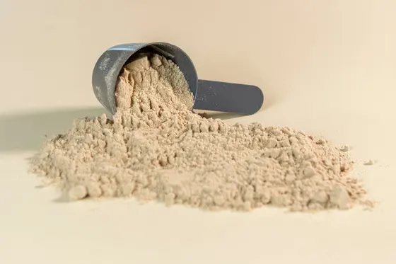 A grey measuring scoop tipped on its side, with light brown protein powder spilling out of it into a heap on a cream surface