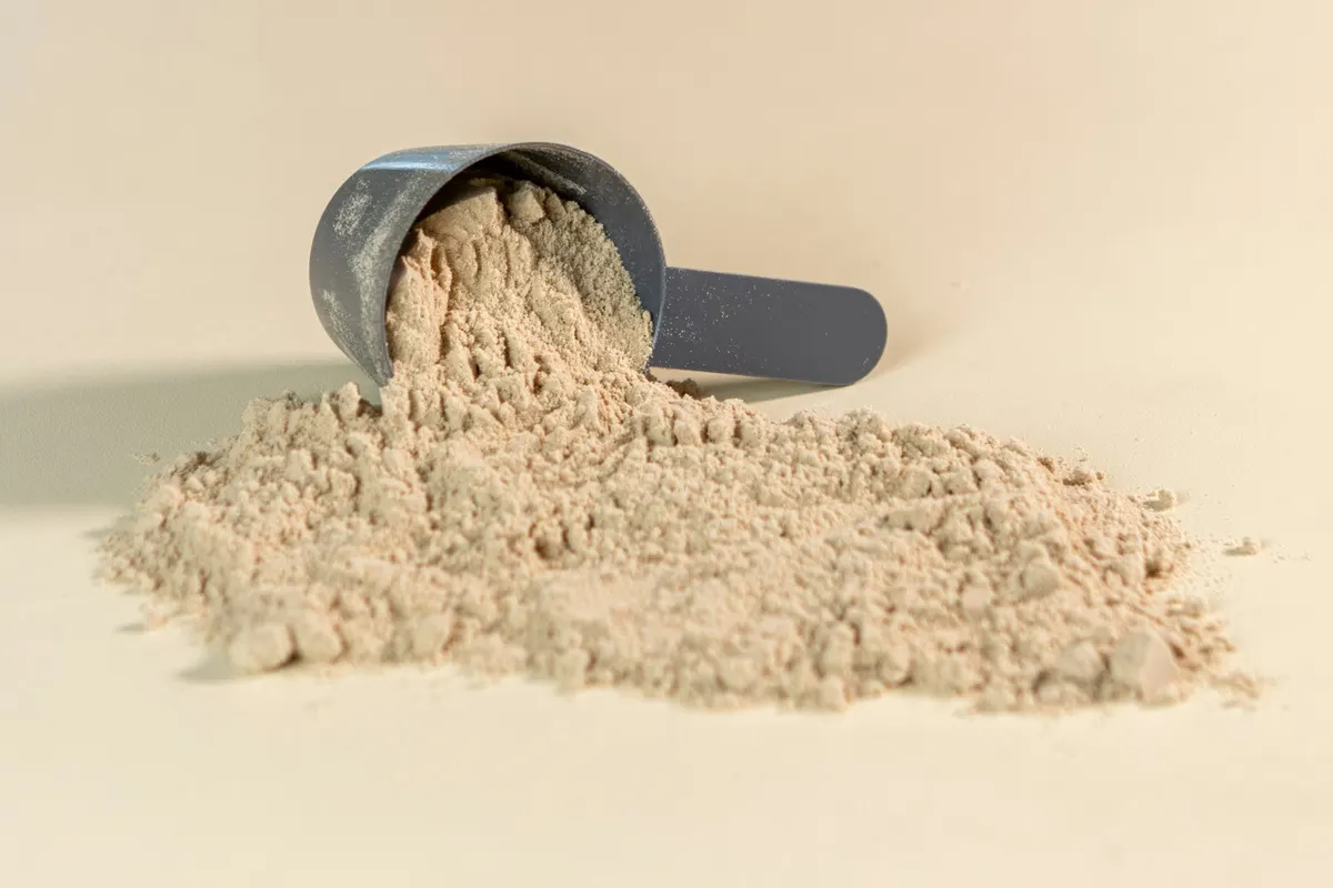 A grey measuring scoop tipped on its side, with light brown protein powder spilling out of it into a heap on a cream surface