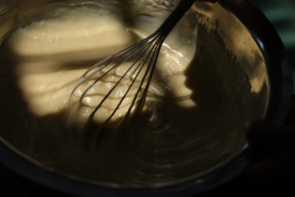 A balloon whisk drawing thick pale sauce up the side of a metal mixing bowl