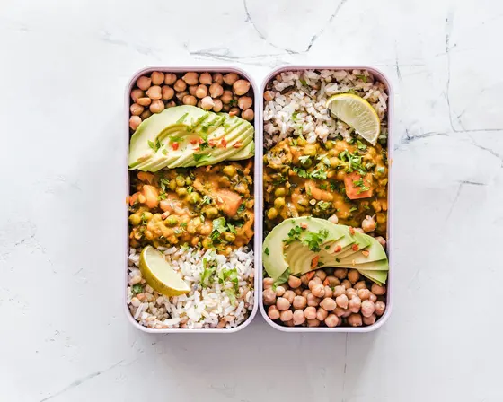 Two lunch boxes side by side on white marble, each holding rice, chickpeas, a vegetable curry, sliced avocado and a wedge of lime
