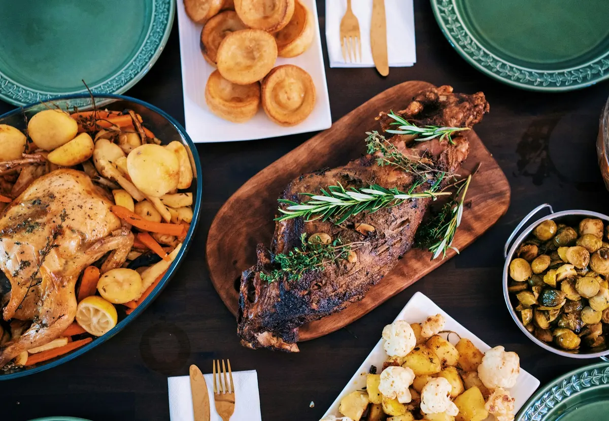 A roast dinner spread across a dark table: a herb-crusted joint on a wooden board, a whole roast chicken with carrots and parsnips, a plate of Yorkshire puddings, sprouts and roast potatoes