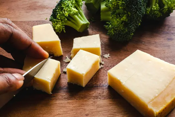 A hand cutting a block of pale cheese into cubes on a wooden board, with broccoli florets behind it