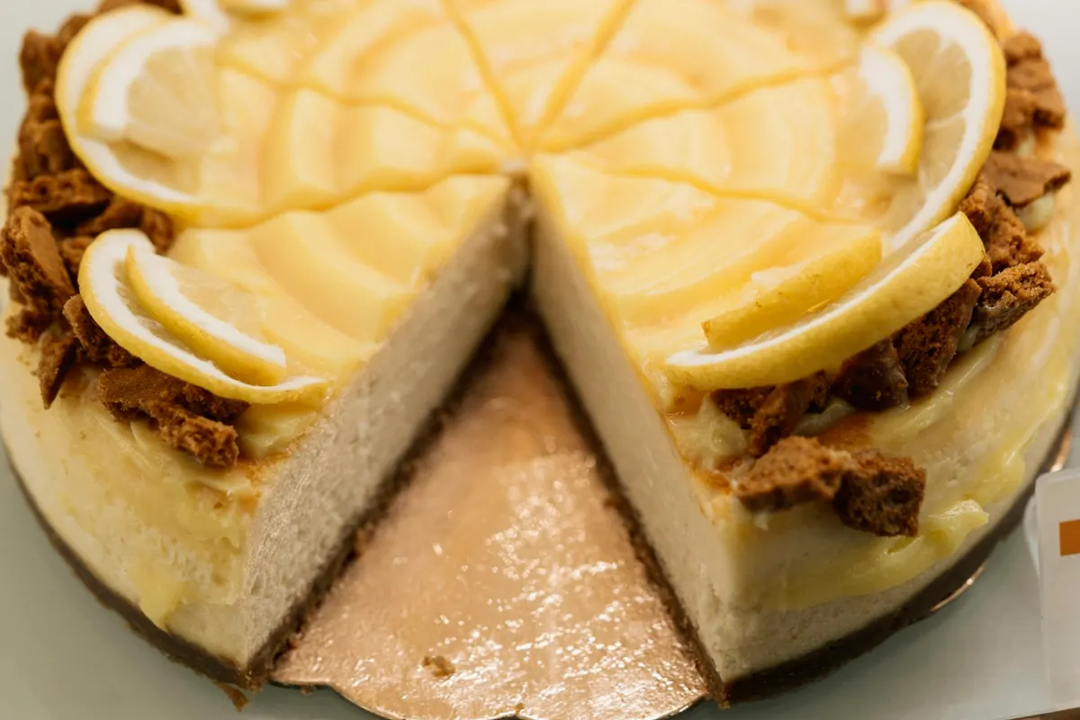 A baked cheesecake with one wedge cut out, showing a pale, even filling on a thin biscuit base, topped with lemon curd, half-slices of lemon and broken spiced biscuits