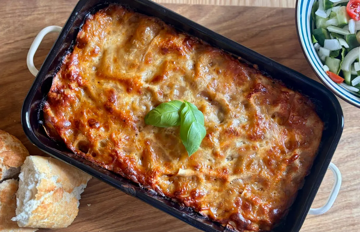 A whole baked lasagna in a black enamel dish, the cheese browned in patches, with a sprig of basil on top and torn bread and a bowl of salad beside it