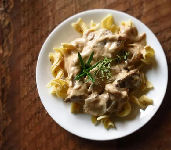 Beef stroganoff with mushrooms in a creamy sauce over egg noodles, with a sprig of fresh herbs, on a white plate