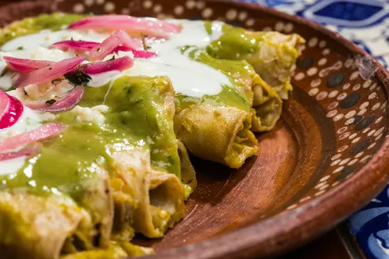 Rolled tortillas under green salsa, crema and pink pickled onion on a clay plate