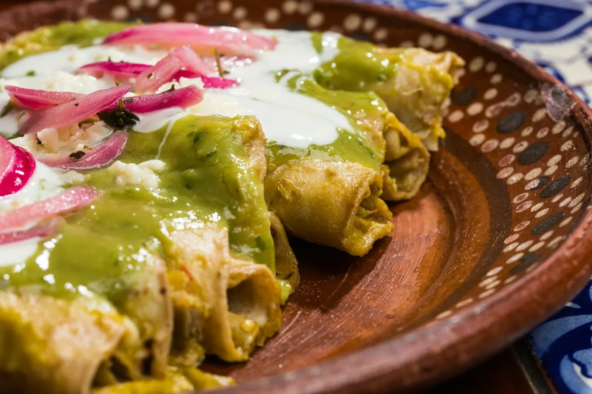 Rolled tortillas under green salsa, crema and pink pickled onion on a clay plate