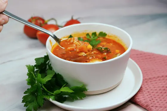 A white bowl of chunky tomato soup with a spoon lifting a mouthful, parsley on top and beside it, and cherry tomatoes on the vine behind