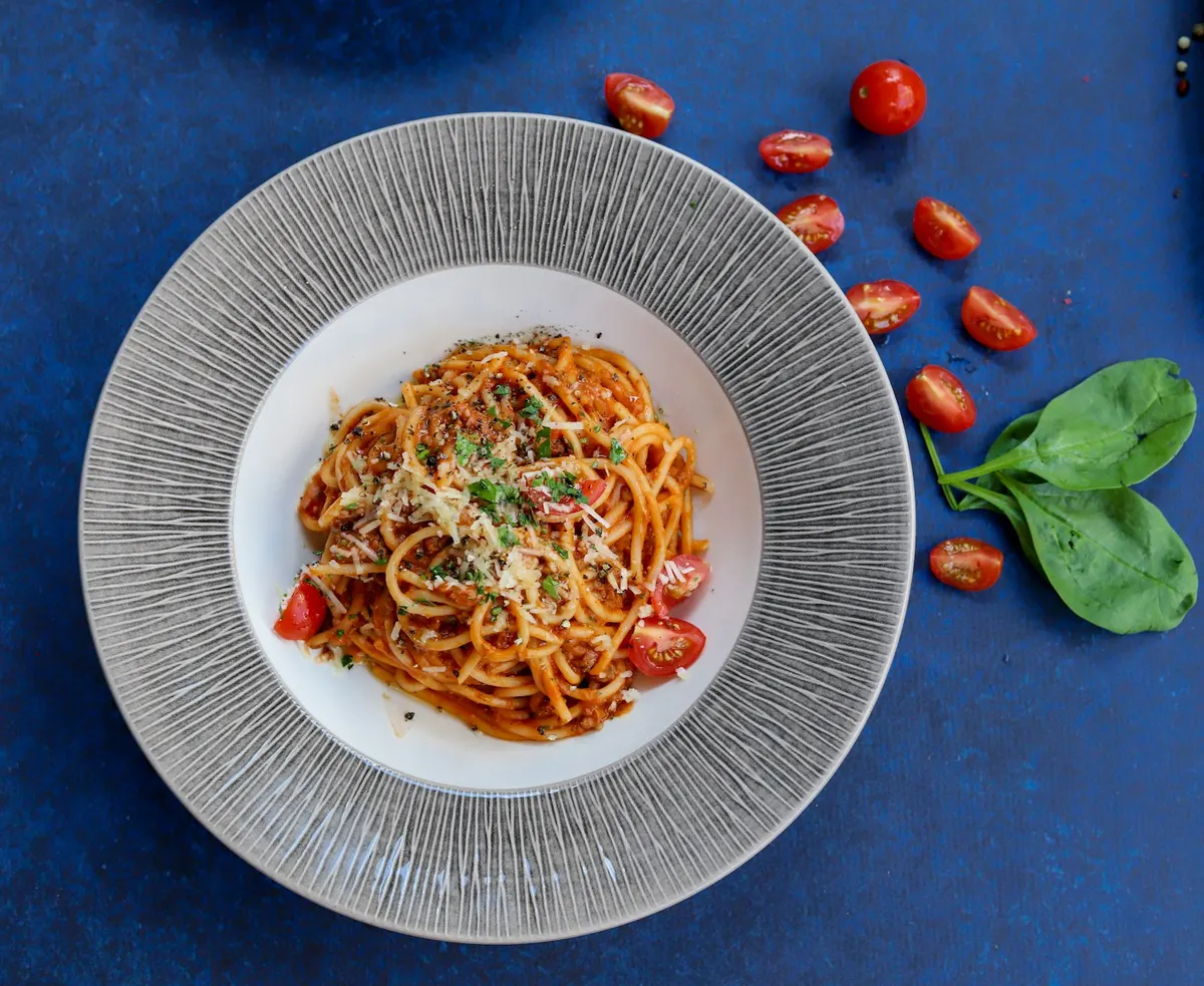 A bowl of spaghetti in tomato sauce topped with grated cheese and chopped herbs, with cherry tomatoes and basil leaves scattered on the surface beside it