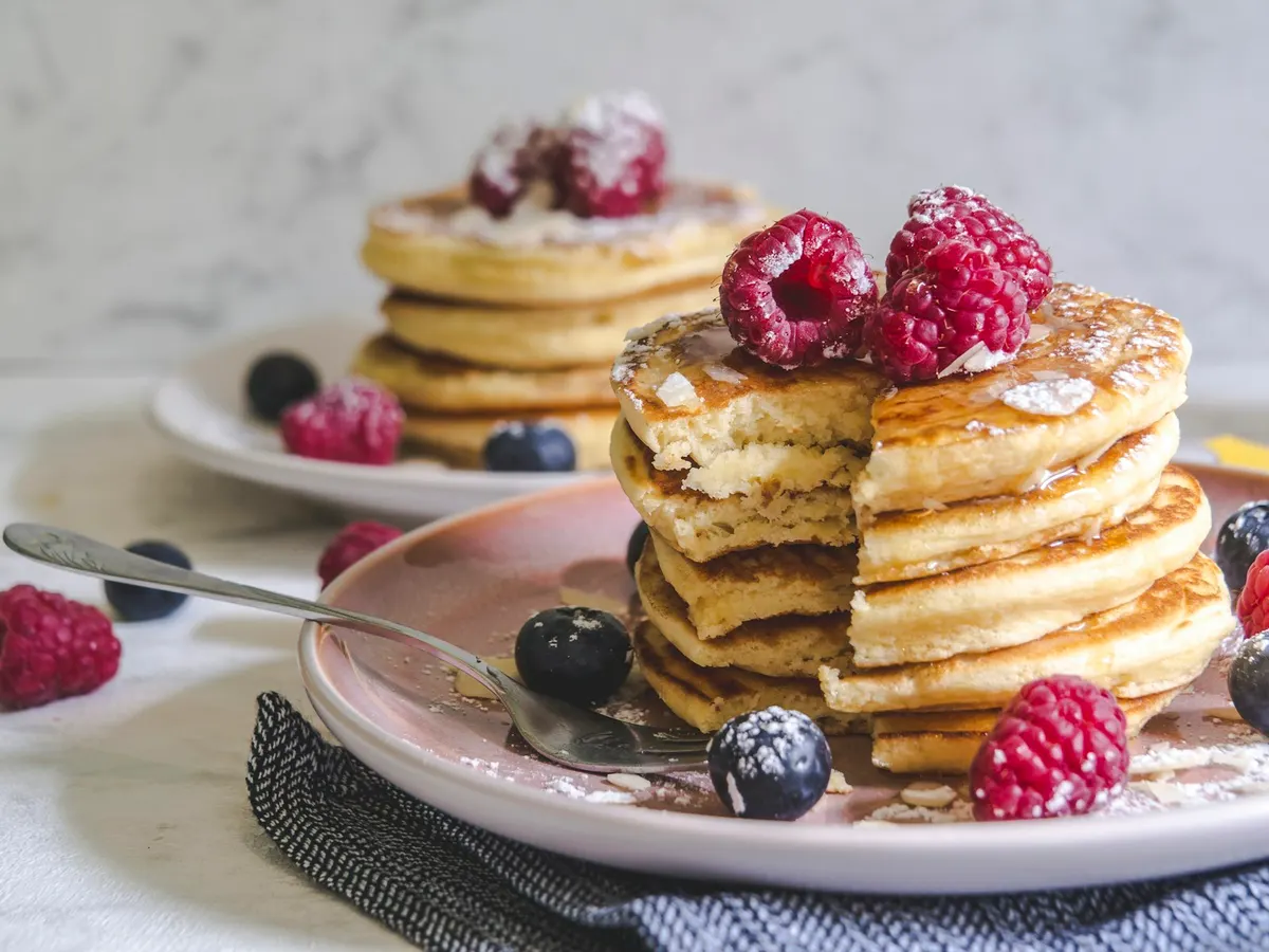 A stack of thick pancakes on a pink plate with a wedge cut away to show the soft crumb, topped with raspberries and a drizzle of syrup, with blueberries and a fork beside it and a second stack behind