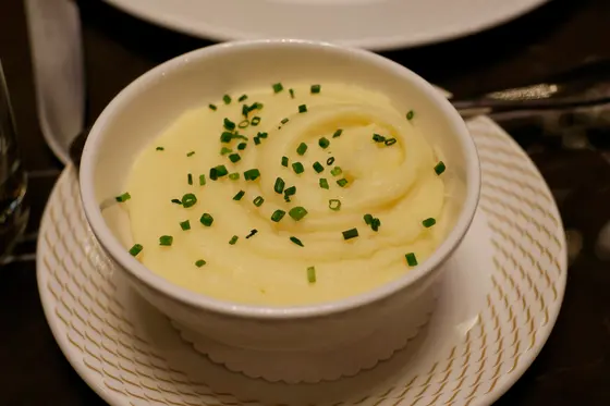 A white bowl of smooth mashed potato swirled into ridges and scattered with chopped chives, on a saucer