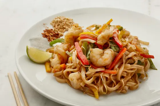 A plate of flat rice noodles with prawns and strips of red and yellow pepper, a lime wedge, crushed peanuts and chilli flakes set beside them