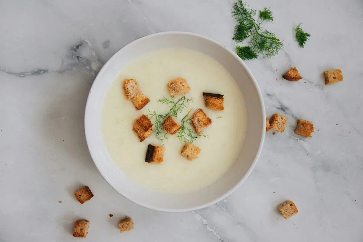 A bowl of smooth, pale creamy soup topped with croutons and sprigs of dill, on a white marble table