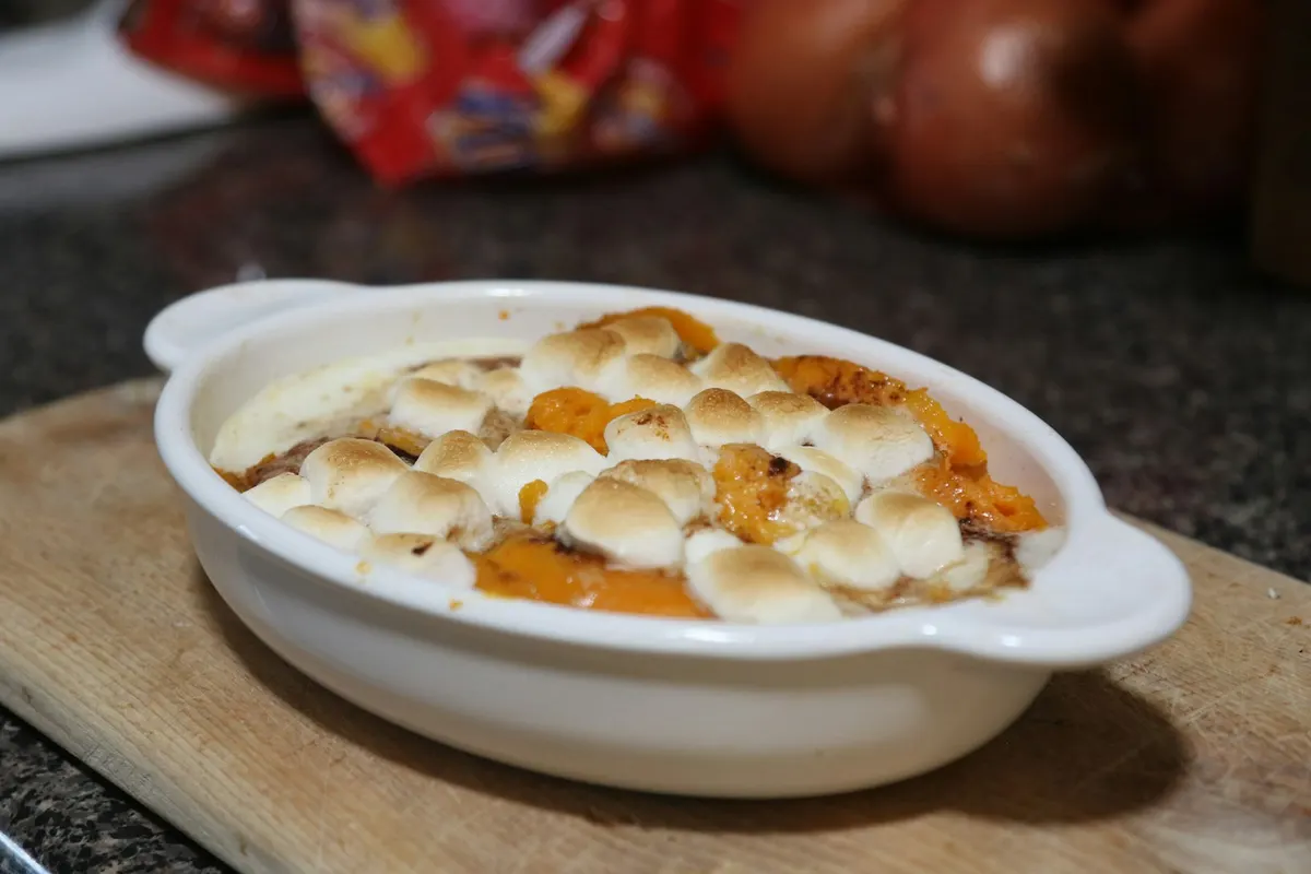 A small oval white dish of sweet potato casserole topped with toasted mini marshmallows, on a wooden board