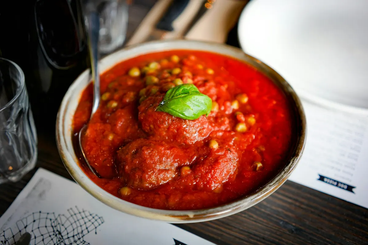 Meatballs in a thick tomato sauce with peas and a basil leaf, served in a stoneware bowl with a spoon