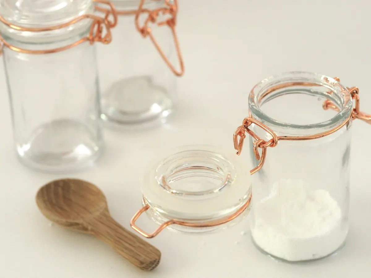 An open clip-top glass jar holding a little white powder beside a small wooden scoop, with two empty jars behind
