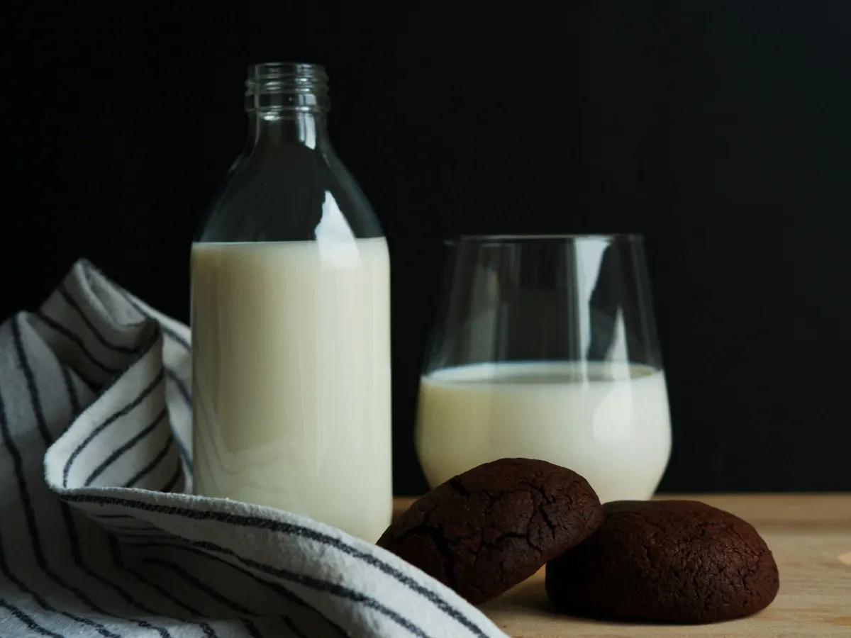 A glass bottle and a glass of milk on a wooden board beside two chocolate cookies, against a dark background