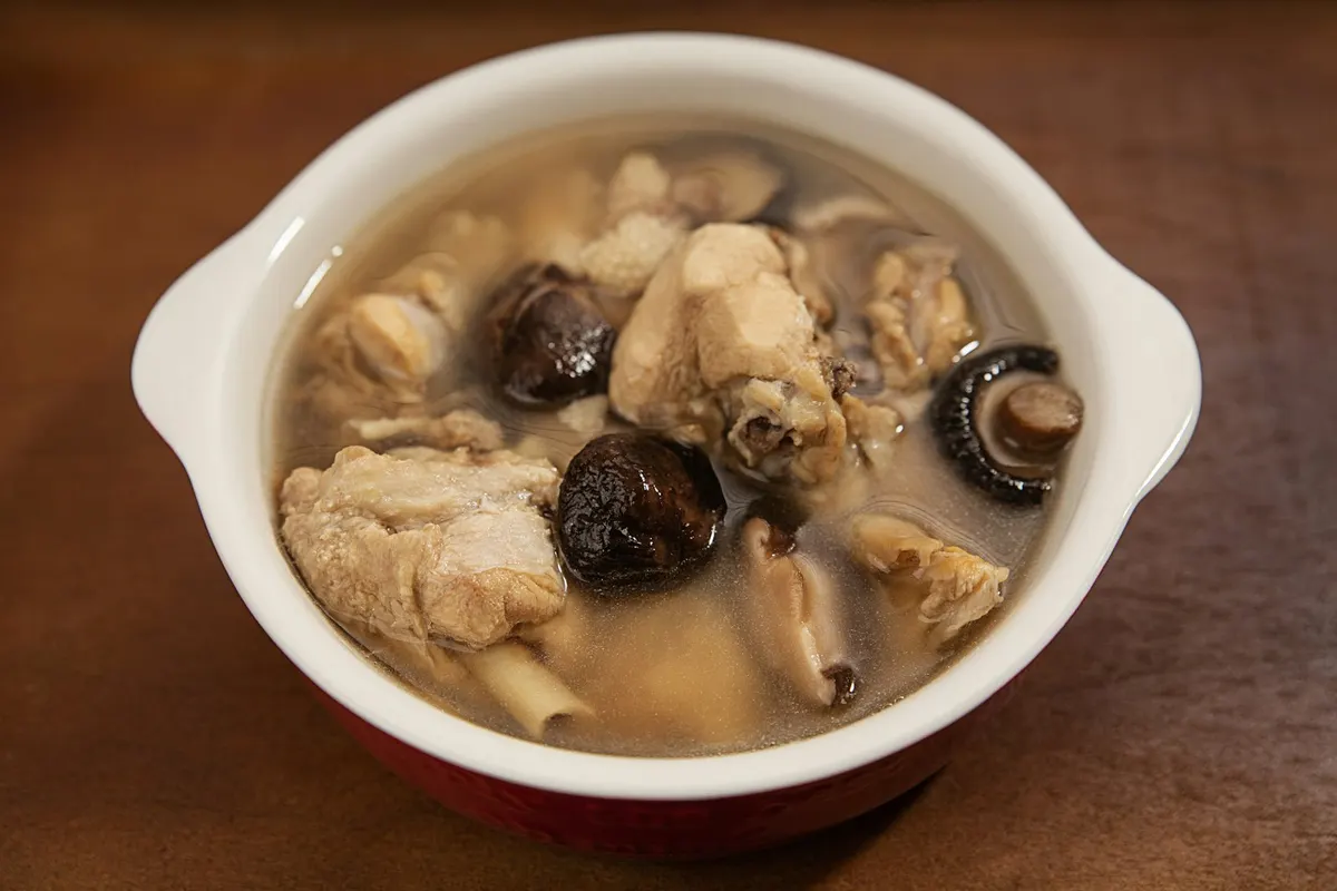 A red and white bowl of clear broth with pieces of chicken on the bone and whole shiitake mushrooms