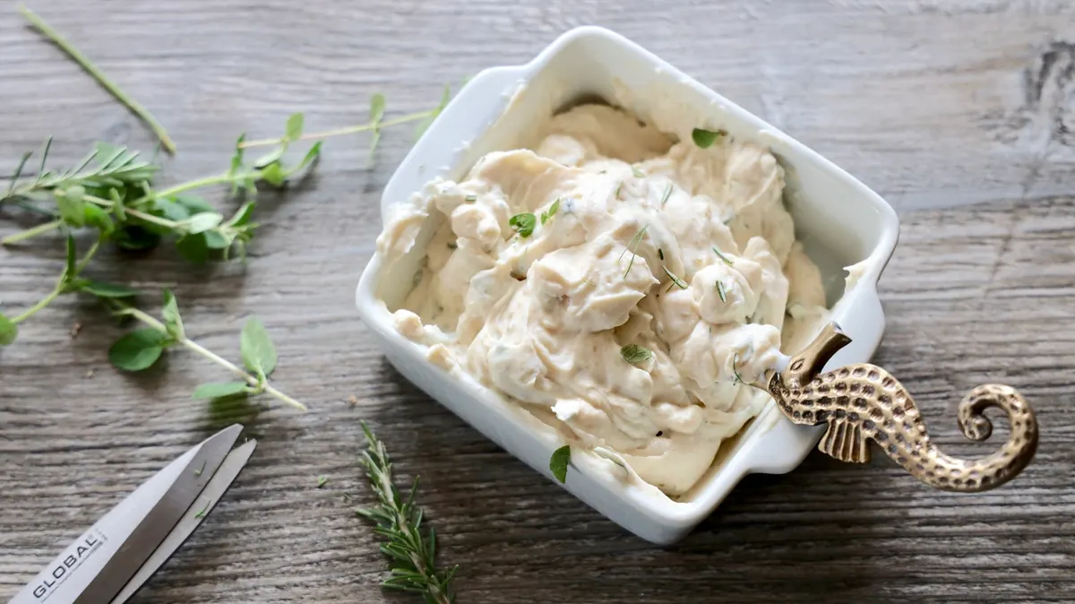 A square white dish of thick soft cheese flecked with chopped herbs, beside sprigs of rosemary and oregano on a weathered wooden board