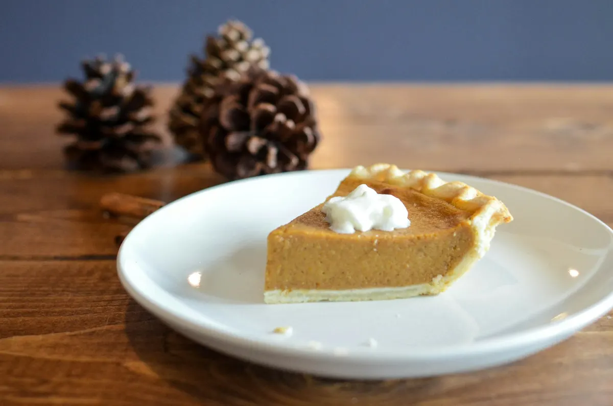 A slice of pumpkin pie with a spoonful of whipped cream on a white plate, on a wooden table with pine cones and a cinnamon stick behind