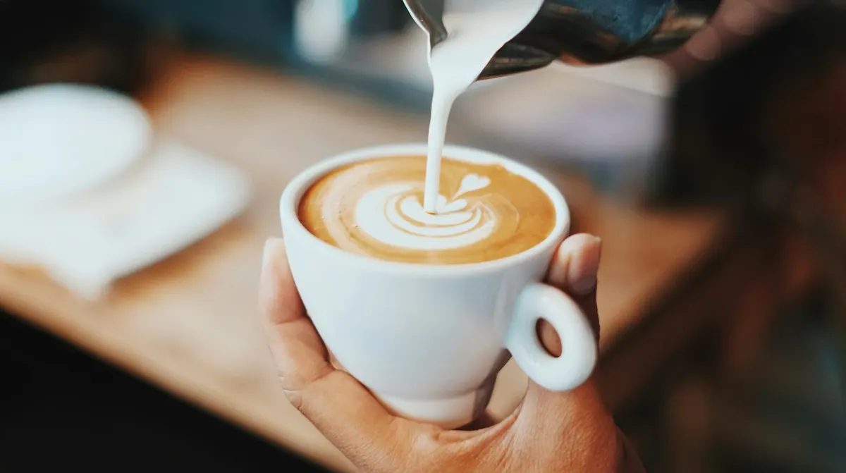A small steel jug pouring a thin stream of milk into a white cup of coffee held in one hand, making a heart pattern in the crema