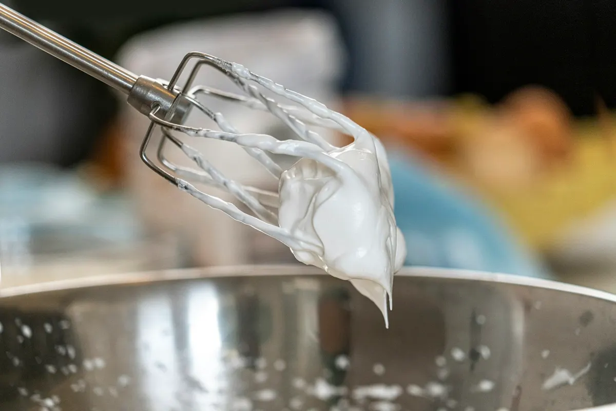 A mixer beater lifted above a steel bowl, holding a thick stiff peak of whipped cream