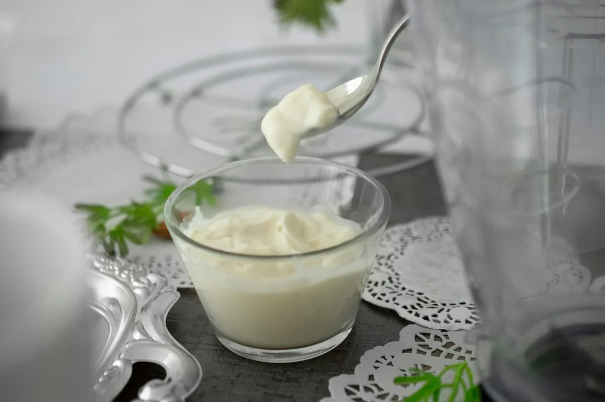A spoon lifting a scoop of thick pale mayonnaise from a small glass bowl