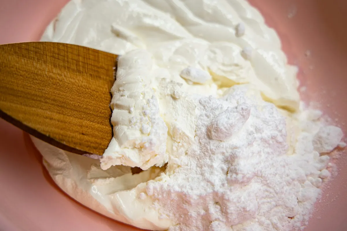 A wooden spatula folding a mound of powdered sugar into cream cheese in a pink bowl