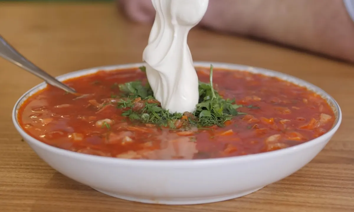 A thick ribbon of sour cream being spooned onto a bowl of red beetroot soup topped with chopped dill and parsley