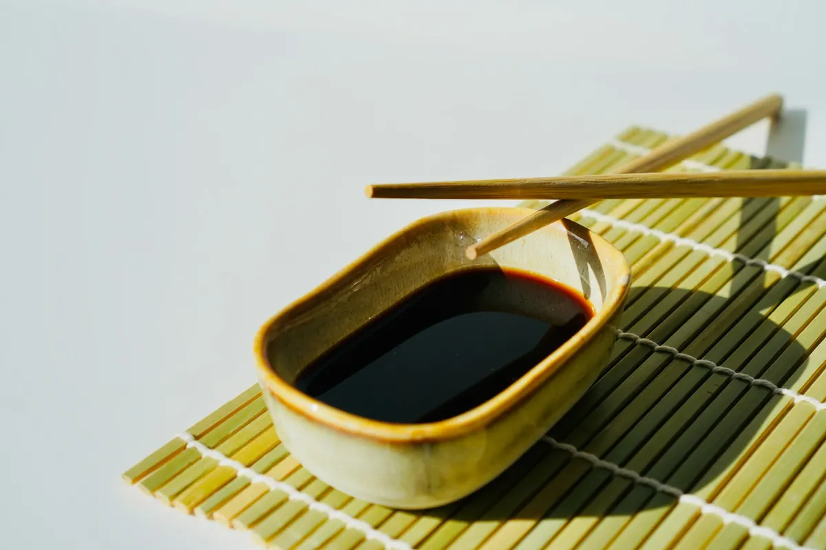 A glazed ceramic dish of dark sauce with chopsticks resting across it, on a bamboo mat