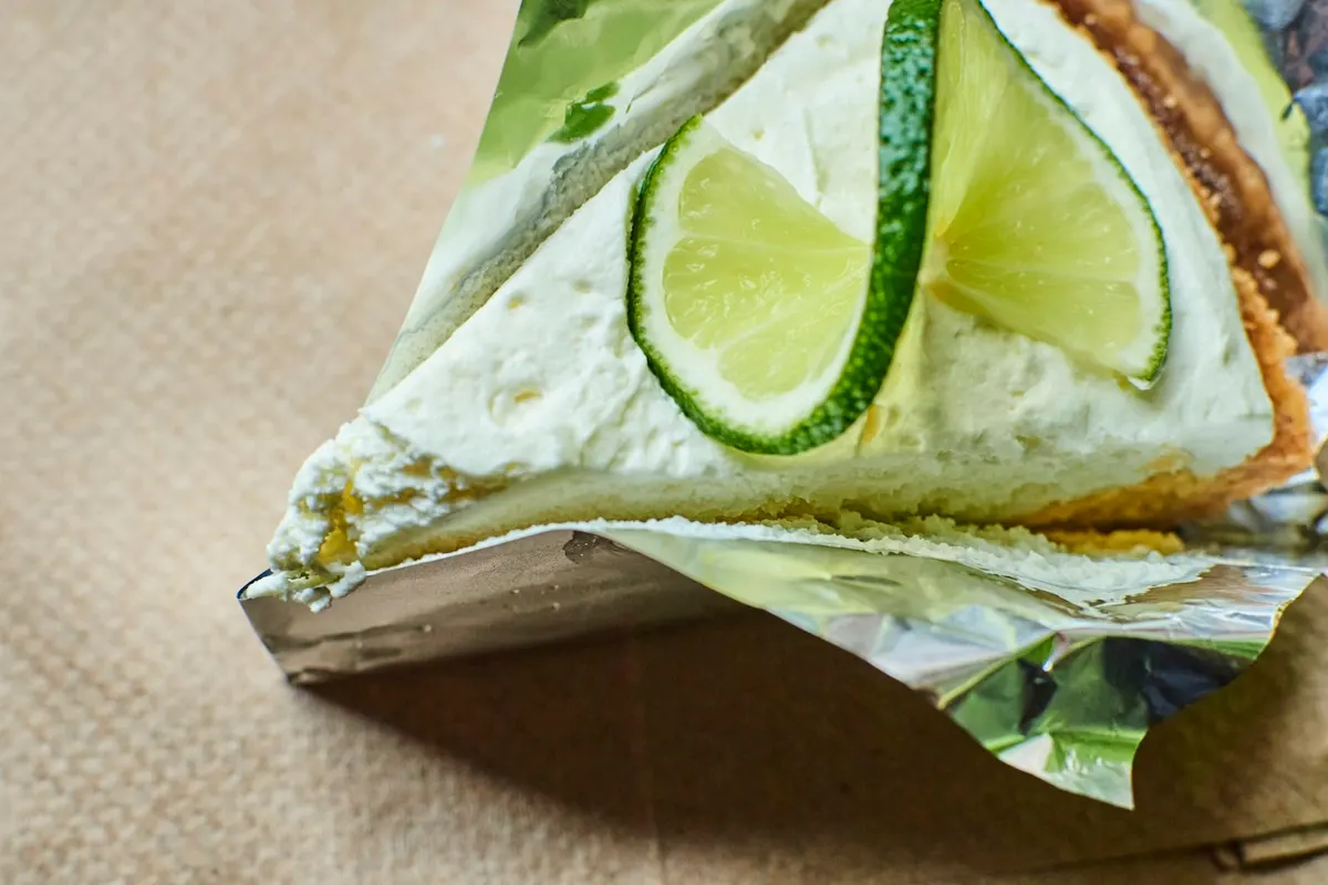 A slice of key lime pie on crumpled foil, topped with whipped cream and two thin lime slices, the biscuit crust visible along one edge