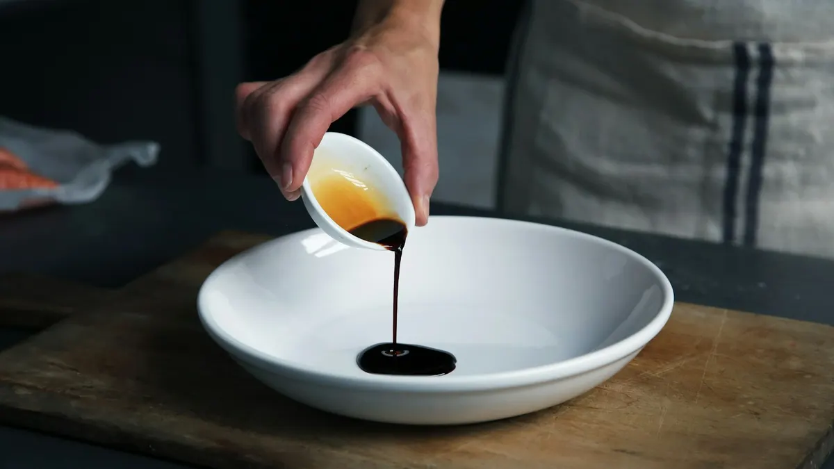 A hand pours a thin stream of dark sauce from a small cup into a white bowl on a wooden board